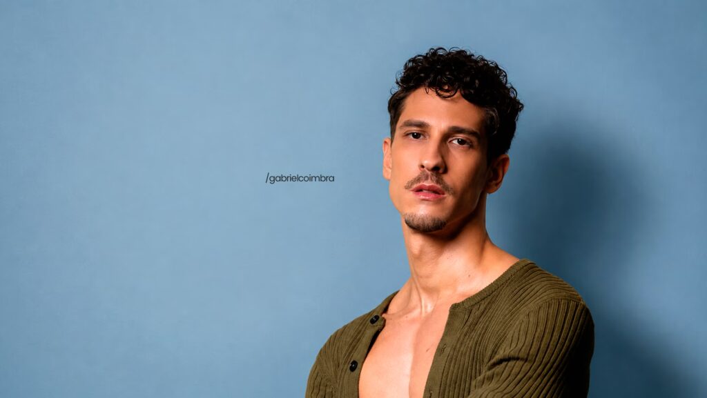 Gabriel Coimbra usando uma camisa verdade que está aberta. Ele olha de forma séria para a frente.