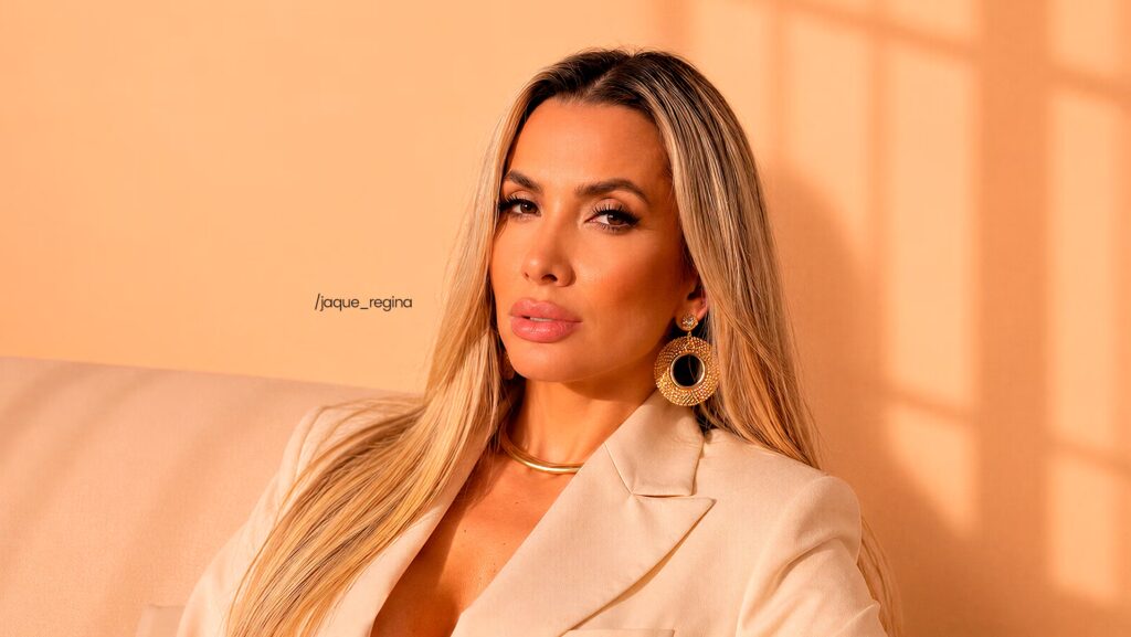 Jaque Regina está sentada. Ela está utilizando uma paletó branco enquanto olha para frente.