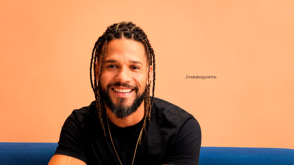 Juninho olhando para a frente e sorrindo. Ele está usando uma camiseta preta.