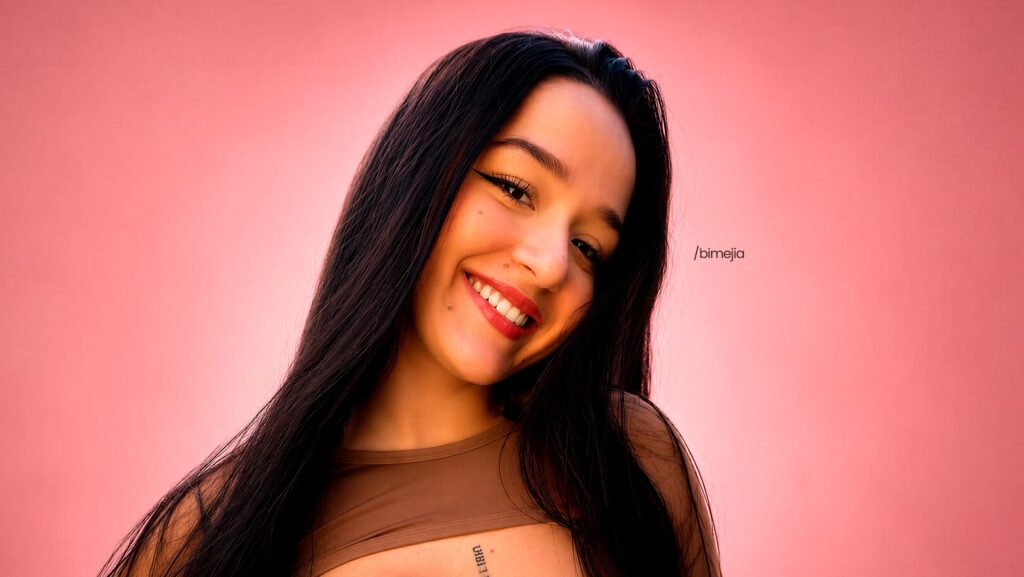 Bi Mejia está sorrindo. Com a cabeça levemente inclinada para a direita.