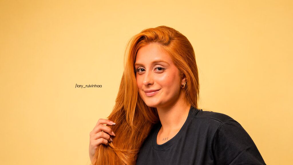 Lary Nascimento com a mão direita no cabelo enquanto solta um sorriso.