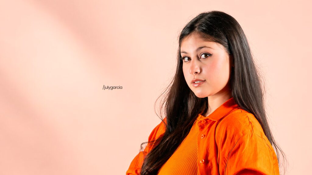 July Garcia está mais a direita da imagem. Utilizando uma roupa laranja e olhando para a imagem.