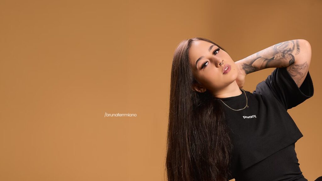 Bruna Fermiano está mais ao lado direito da imagem. Ela está sentada com sua mão na nuca. Ao fundo, uma parede bege.