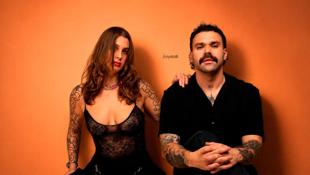 foto de Casal Crystalli encostado em uma parede. Ambos estão usando roupas pretas. A parede é da cor laranja.