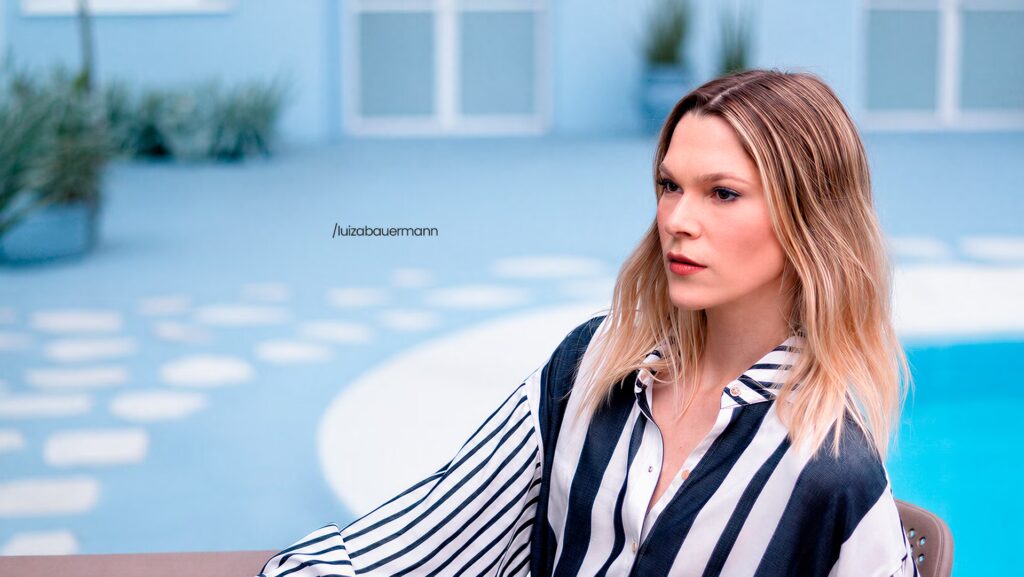 Luiza Bauermann está usando uma camisa com listras verticais pretas e brancas. Ela está olhando de lado. Em um pose para foto.