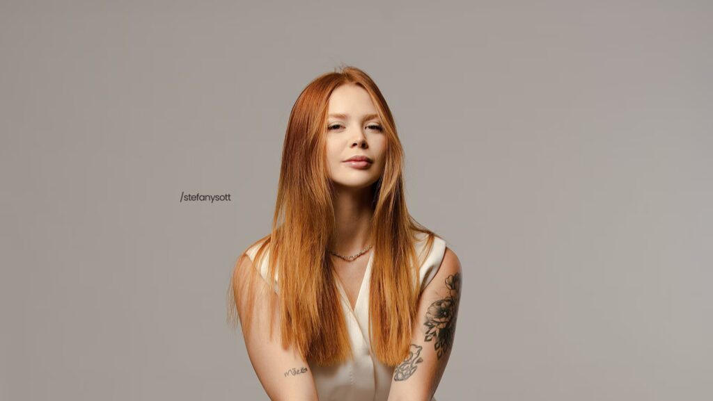 Stefany Sott está olhando séria para a imagem. Ela está utilizando um vestido branco e a parede de fundo é cinza.