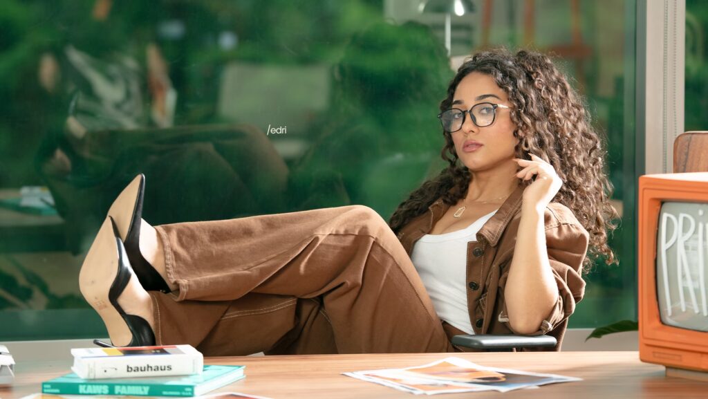 Edriane Dantas está sentada numa cadeira com os pés em cima de uma mesa. Ela usa uma calça marrom