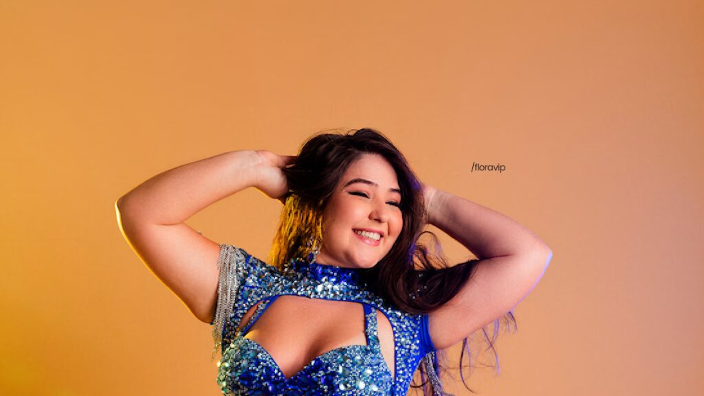 Flora Favaretto olha de forma sorridente para o lado enquanto está com as duas mãos na cabeça. Ela usa um vestido azul e o fundo da imagem é uma parede laranja.