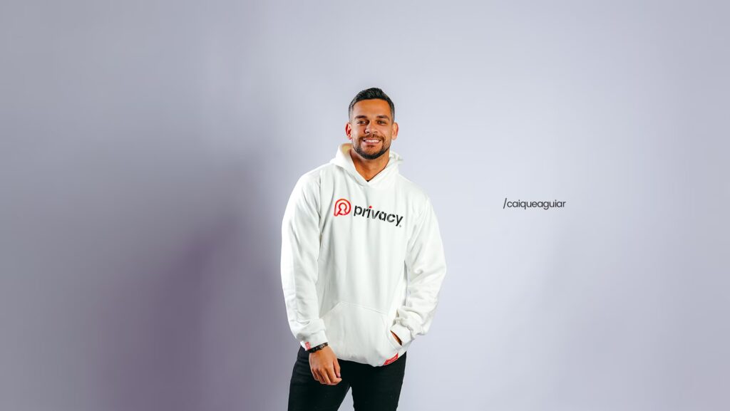Caique Aguiar sorrindo. Ele está usando um moletom branco.