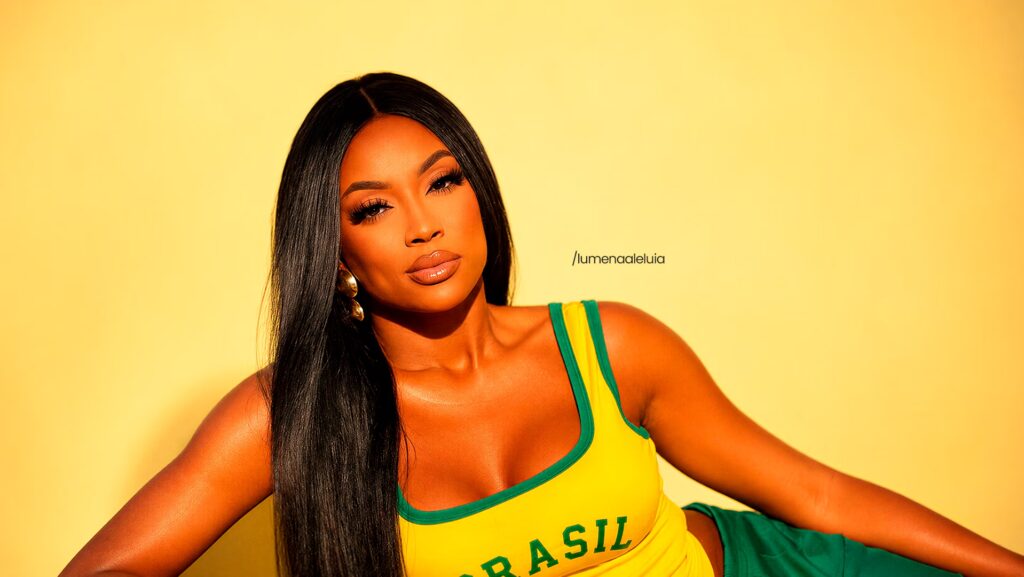 Lumena está sentada e olhando para frente. Ela usa uma blusinha amarela do Brasil.